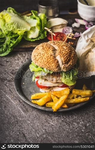 Homemade tasty burger with chicken, lettuce, mozzarella and tomatoes served with french fries potatoes on rustic kitchen table background, front view