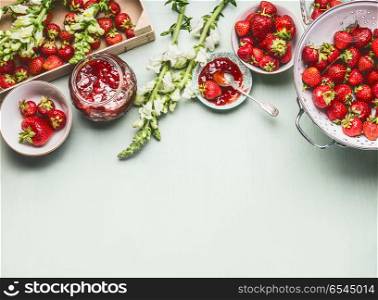 Homemade tasty strawberries jam in glass jar with summer flowers and fresh berries, bowls and spoon on table background, top view. Berries preserve concept, border