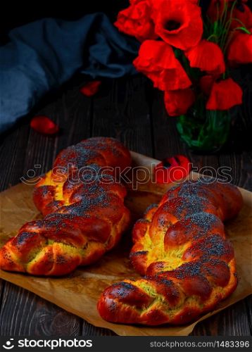 Homemade wicker challah for Shabbat. Homemade sweet Finnish Pulla or Zopf bread with poppy seeds and saffron on a baking sheet, close up. Blooming poppies on a wooden table.