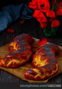 Homemade wicker challah for Shabbat. Homemade sweet Finnish Pulla or Zopf bread with poppy seeds and saffron on a baking sheet, close up. Blooming poppies on a wooden table.