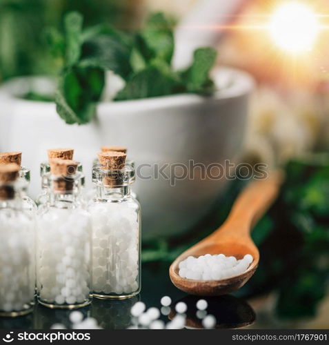 Homeopathy Concept. Homeopathic globules spilled around, wooden spoon filled with homeopathic pills, decorated background with fresh green herbs and flowers. . Homeopathy Concept