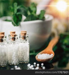 Homeopathy Concept. Homeopathic globules spilled around, wooden spoon filled with homeopathic pills, decorated background with fresh green herbs and flowers. . Homeopathy Concept