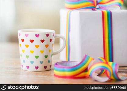 homosexual and lgbt concept - close up of coffee cup with gift box and gay pride awareness ribbon on wooden table at home. close up of cup, gift box and gay awareness ribbon. close up of cup, gift box and gay awareness ribbon
