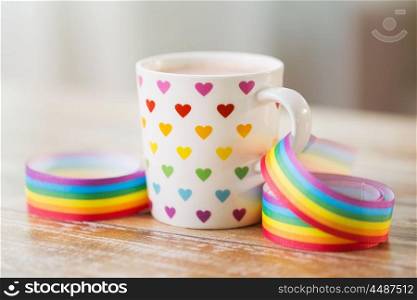 homosexual and lgbt concept - close up of cup with rainbow colored heart pattern and gay pride awareness ribbon on wooden table. cup with heart pattern and gay awareness ribbon. cup with heart pattern and gay awareness ribbon