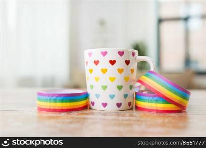 homosexual and lgbt concept - close up of cup with rainbow colored heart pattern and gay pride awareness ribbon on wooden table at home. cup with heart pattern and gay awareness ribbon. cup with heart pattern and gay awareness ribbon