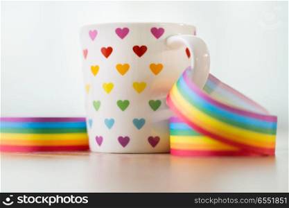 homosexual and lgbt concept - close up of cup with rainbow colored heart pattern and gay pride awareness ribbon on wooden table. cup with heart pattern and gay awareness ribbon. cup with heart pattern and gay awareness ribbon