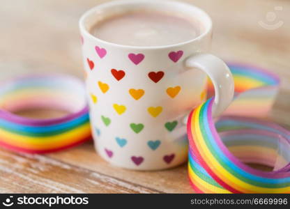 homosexual and lgbt concept - close up of cup with rainbow colored heart pattern and gay pride awareness ribbon on wooden table. cup with heart pattern and gay awareness ribbon. cup with heart pattern and gay awareness ribbon