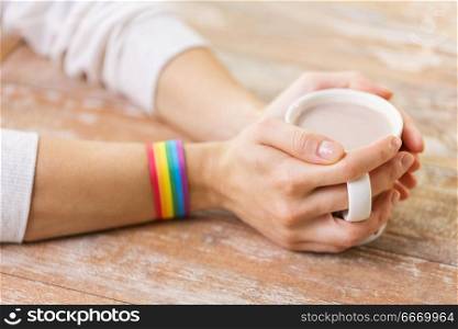 homosexual and lgbt concept - close up of female hand with cacao drink in cup with heart pattern and gay pride awareness wristband on wooden table. hands with cacao cup and gay awareness wristband. hands with cacao cup and gay awareness wristband