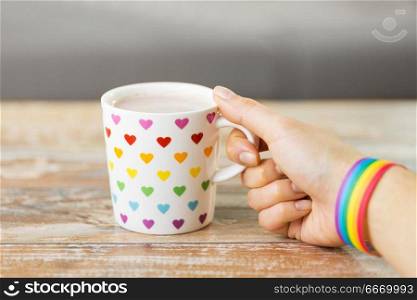 homosexual and lgbt concept - close up of female hand with cacao drink in cup with heart pattern and gay pride awareness wristband on wooden table. hand with cup of cacao and gay awareness wristband. hand with cup of cacao and gay awareness wristband