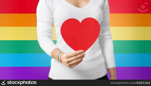 homosexual and lgbt concept - woman holding red heart shape and wearing gay pride awareness ribbon wristband over rainbow background. woman with gay awareness wristband holding heart