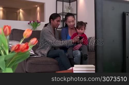 Homosexual couple, gay people, young lesbian women, same sex marriage relationship between Asian girls. Multi-ethnic family watching TV with adopted child, mixed race lesbians having fun with baby