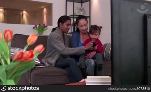 Homosexual couple, gay people, young lesbian women, same sex marriage relationship between Asian girls. Multi-ethnic family watching TV with adopted child, mixed race lesbians having fun with baby
