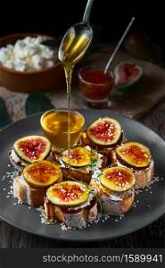 Honey dripping from a spoon onto fig sandwiches on toasted goat cheese baguette on a plate with honey, sesame seeds and walnuts. Fresh toast with figs and garnished with thyme leaves. Close-up