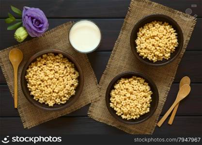 Honey flavored breakfast cereal in three rustic bowls with a glass of milk and wooden spoons on the side, photographed overhead on dark wood with natural light