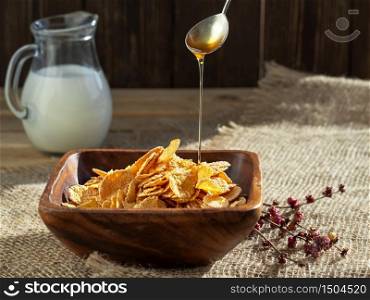 Honey from a spoon is poured into corn cereal. Jug of milk , corn cereals in a wooden plate on dark wooden table. Closeup, sunlight. Shallow depth of field.
