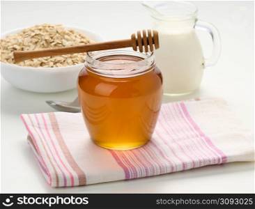 honey in a glass transparent jar and a wooden stick on a white table, behind a decanter with milk and oatmeal in a plate