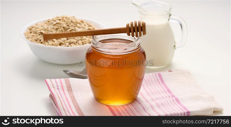 honey in a glass transparent jar and a wooden stick on a white table, behind a decanter with milk and oatmeal in a plate