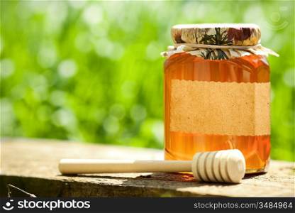 Honey jar with blank paper label and wooden stick on table against green spring natural background