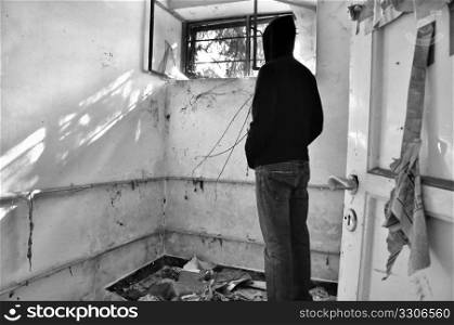 Hooded male figure looking through window of abandoned house. Black and white.