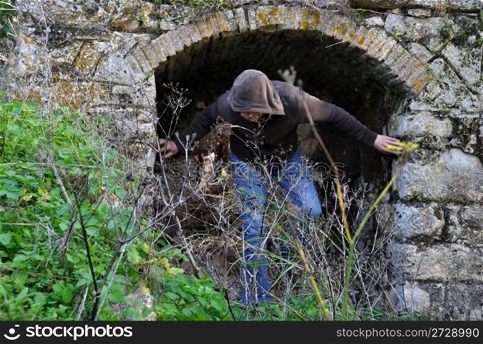 Hooded man obscured by overgrown plants under arched recess of rural ruin.