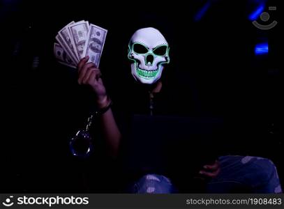 Hoodie male hacker with skull mask holding laptop and money. Horror, Halloween, Technology and Crime Concept.