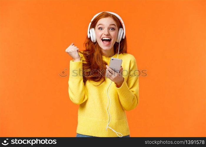 Hooray yes new song. Attractive cheerful and excited redhead woman fist pump in joy and positive emotions, wearing headphones, holding smartphone, smiling camera happily, orange background.. Hooray yes new song. Attractive cheerful and excited redhead woman fist pump in joy and positive emotions, wearing headphones, holding smartphone, smiling camera happily, orange background