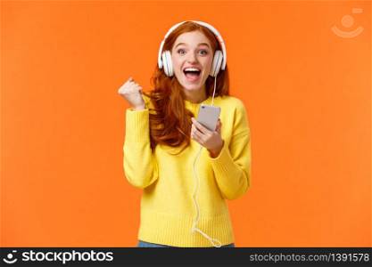 Hooray yes new song. Attractive cheerful and excited redhead woman fist pump in joy and positive emotions, wearing headphones, holding smartphone, smiling camera happily, orange background.. Hooray yes new song. Attractive cheerful and excited redhead woman fist pump in joy and positive emotions, wearing headphones, holding smartphone, smiling camera happily, orange background