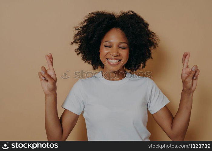 Hopeful 20s dark skinned woman with closed eyes wishing for good luck isolated on beige studio background crossing fingers and making wish, hoping for win and dream come true. Hopeful dark skinned woman with closed eyes wishing for luck isolated on beige studio background