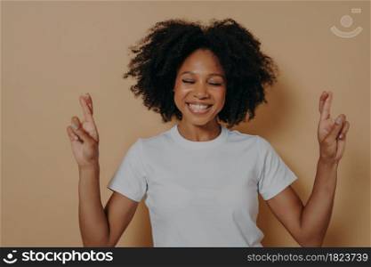 Hopeful 20s dark skinned woman with closed eyes wishing for good luck isolated on beige studio background crossing fingers and making wish, hoping for win and dream come true. Hopeful dark skinned woman with closed eyes wishing for luck isolated on beige studio background