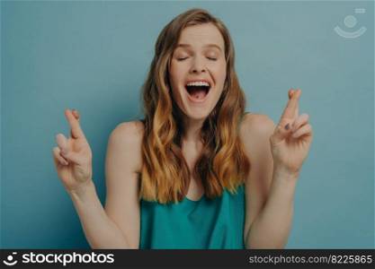 Hopeful emotional woman crossing fingers making wish come true gesture with eyes closed and her mouth wide open from excitation, isolated on blue background. Superstition concept. Hopeful girl crossing fingers making wish come true gesture with eyes closed and her mouth wide open