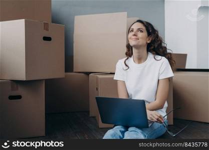 Hopeful hispanic woman sitting among cardboard boxes and holding pc. Lady is in waiting for shipping service and chatting online. Romantic relationship and relocation concept.. Hopeful hispanic woman sitting among cardboard boxes and chatting online. Romantic relationship.