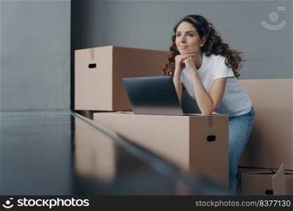 Hopeful spanish woman unpacking cardboard boxes at new place. Lady is looking through window in new apartment. Happy girl in white t-shirt with pc on the box. Relocation and distance work concept.. Hopeful spanish woman unpacking cardboard boxes at new place and looking through window.