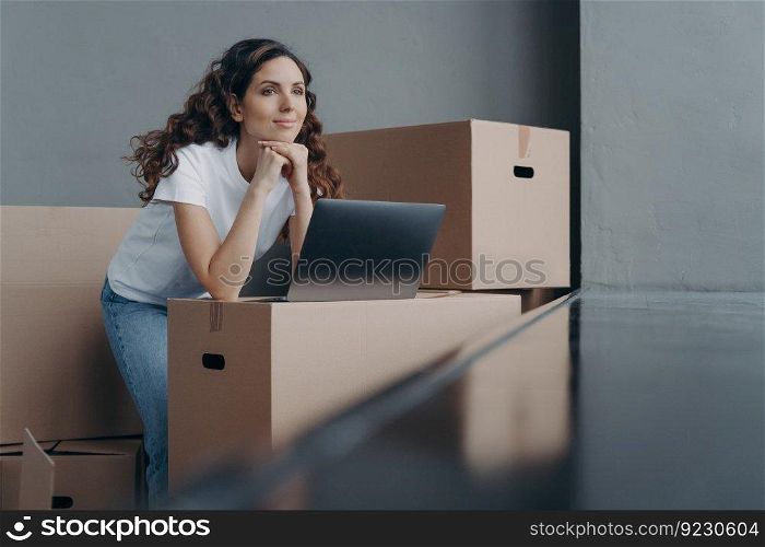 Hopeful spanish woman unpacking cardboard boxes at new place. Lady is looking through window in new apartment. Happy girl in white t-shirt with pc on the box. Relocation and distance work concept.. Hopeful spanish woman unpacking cardboard boxes at new place and looking through window.