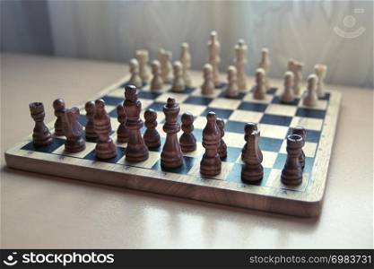 Horizontal close-up side view photographic image of a retro style wooden material chessboard with chess pieces set ready for strategic mind game. Dark brown colored figures in front.