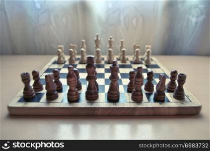 Horizontal close-up side view photographic image of a retro style wooden material chessboard with chess pieces set ready for strategic mind game. Dark brown colored figures in front.