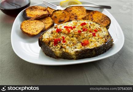 Horizontal closeup photo of baked fish and yam slices on white plate with fork, wine glass, lemon slice and table cloth underneath