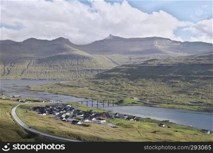 Horizontal colorful scenic image of Faroese landscape with asphalt highway and The bridge between Streymoy and Eysturoy nearby small village with distant mountains in background and white clouds on top. Glorious sceneries of the Faroes. Postcard motif.