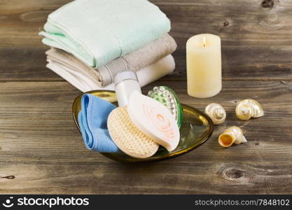 Horizontal front top view of spa hygiene accessories in glass bowl, light white candle with clean stacked towels in background and seashells on rustic wood