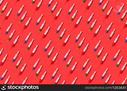 Horizontal health-care pattern from disposable plastic needles with red, green and blue liquid or vaccine on a red background with soft shadow. Flat lay.. Medical pattern of plastic syrenges with multicolor vaccines.