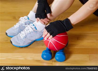 Horizontal image of female hands wearing workout gloves while resting hand on small weight ball with wooden gym floor and partial body in background