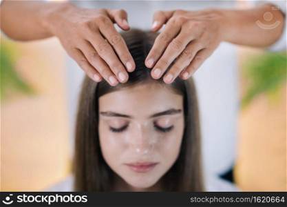 Horizontal image of female Reiki therapist standing and holding hands above head of the beautiful teenage patient. Reiki Practitioner transfers energy and healing crown chakra. Peaceful girl sitting with her eyes closed. Alternative therapy concept. 