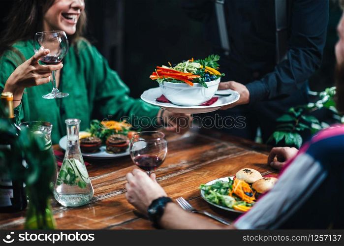 Horizontal image of male hand serving plate of delicious vegetarian mixed organic salad in restaurant to young smiling couple drinking red wine. Waiter wearing black uniform.