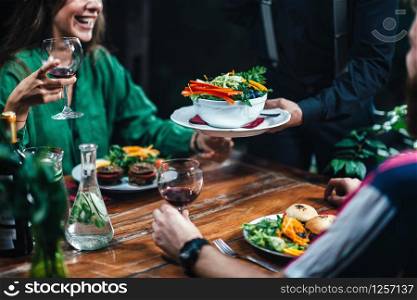 Horizontal image of male hand serving plate of delicious vegetarian mixed organic salad in restaurant to young smiling couple drinking red wine. Waiter wearing black uniform.