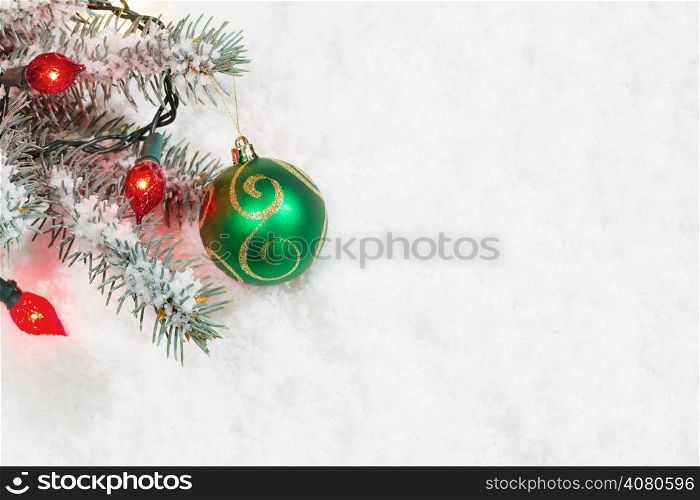 Horizontal image of single Christmas ornament, green and gold, hanging from real Blue Spruce tree branch surrounded by snow and lights