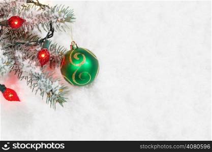 Horizontal image of single Christmas ornament, green and gold, hanging from real Blue Spruce tree branch surrounded by snow and lights