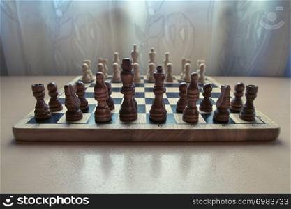 Horizontal low angle view photographic image of a retro style wooden material chessboard with chess pieces set ready for strategic mind game. Dark brown colored figures in front.