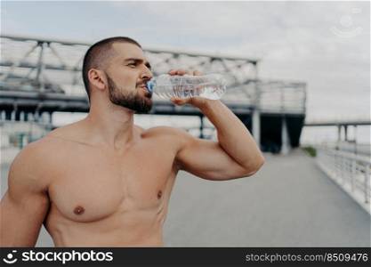 Horizontal outdoor shot of sporty bearded athlete man drinks water after training, holds bottle of refreshing drink, has break during exercising in open air, poses near bridge. Sportsman drinking