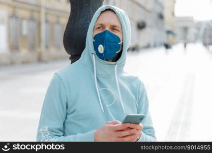 Horizontal outdoor shot of young man wears respirator mask for health protection, walks on street with mobile phone, protects himself during coronavirus outbreak, follow news online, dressed in hoodie