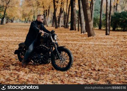 Horizontal outdoor view of active male motorcyclist rides bike, wears trendy sunglasses and black jacket, poses in big autumn park, enjoys awesome travel, has motorcycle trip, rest at nature.