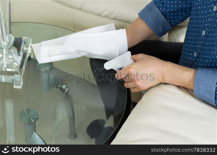 Horizontal photo female hand spraying cleaning solution, from spray bottle, onto dirty glass round end table with paper towels and sofa with partial lamp in background